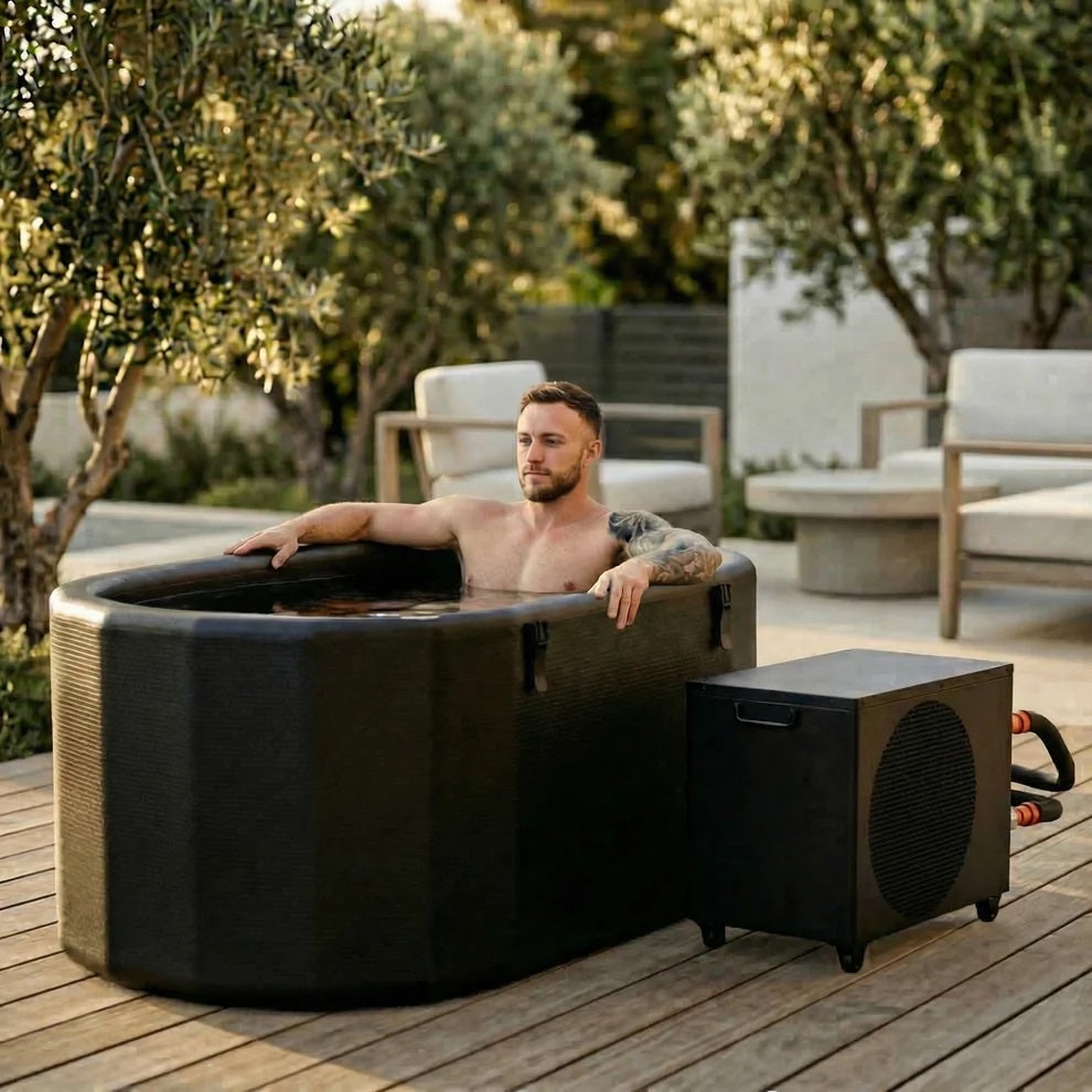 A man relaxing in a black outdoor cold plunge tub with its chiller unit, on a wooden deck surrounded by olive trees.